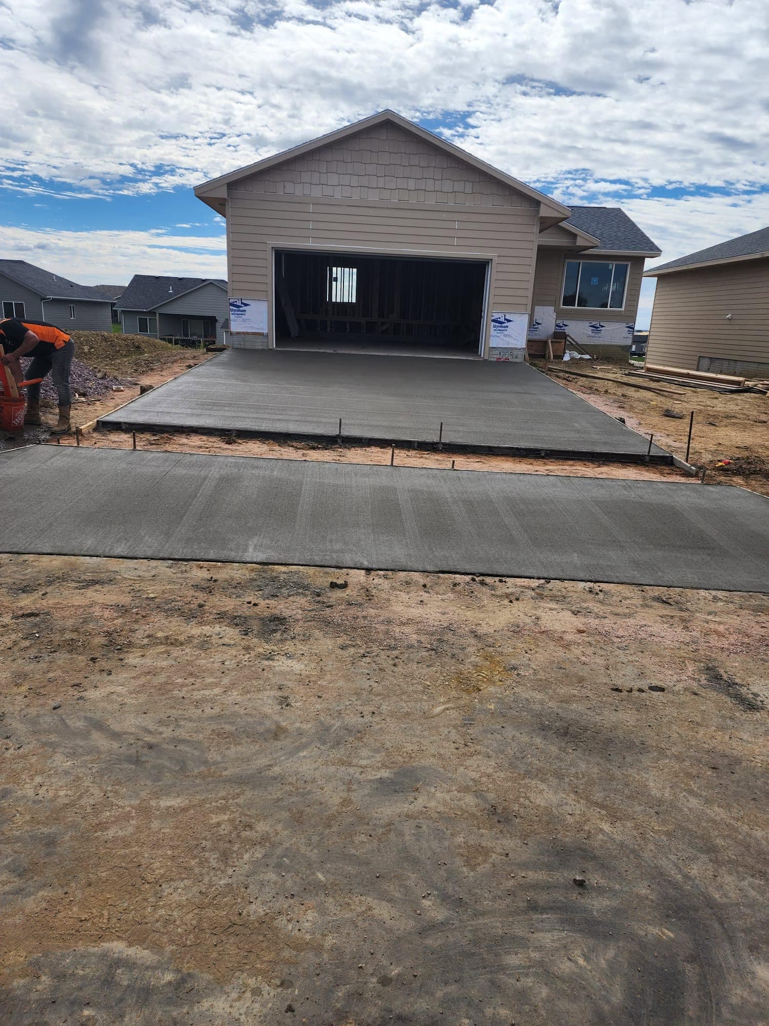 A wide fresh concrete driveway and apron at the open framed garage of a new home, a worker bent over its far left edge