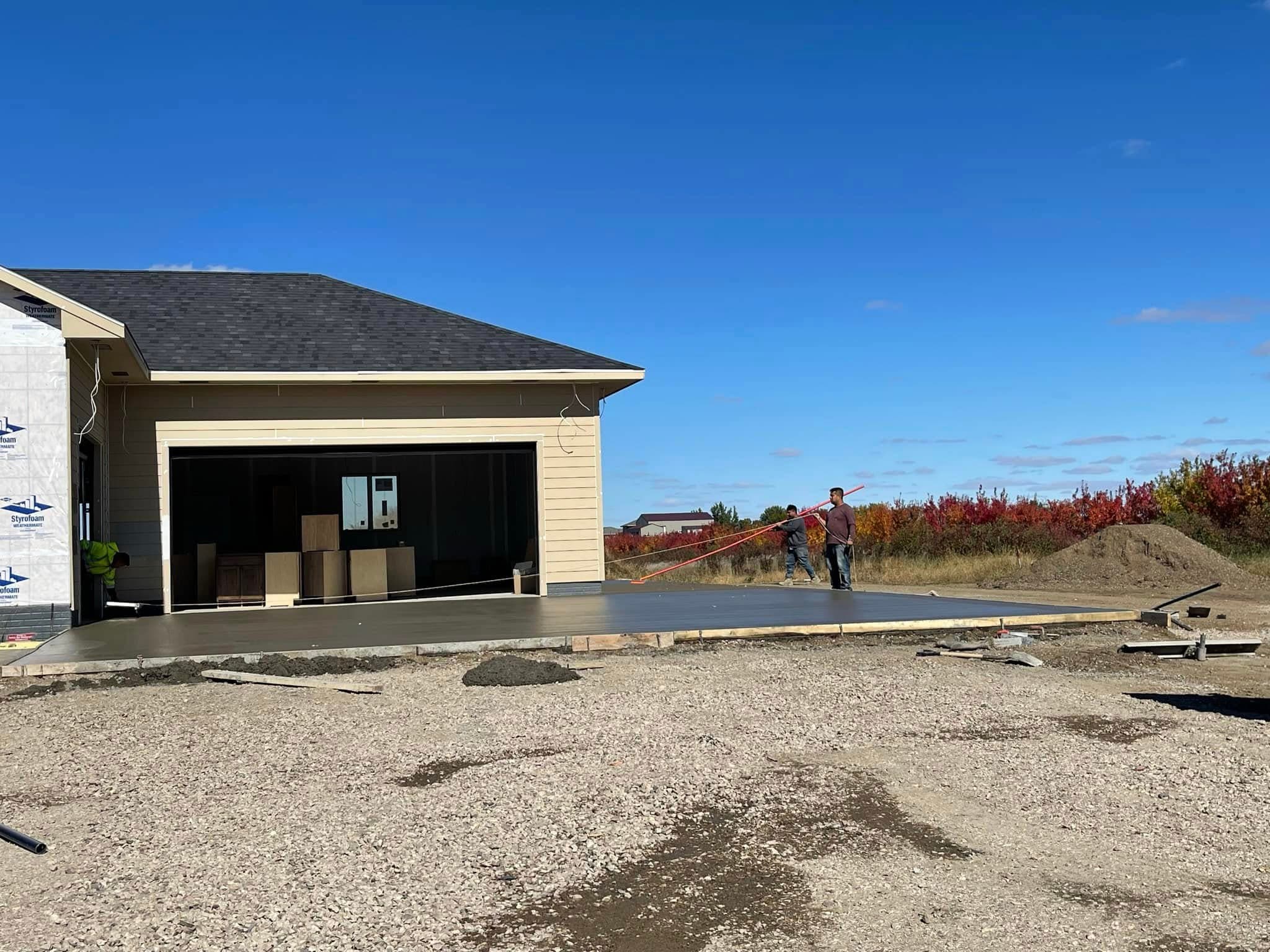 A wet just-poured concrete slab at the open garage of a home under construction, two workers running a long-handled float across it from the far edge