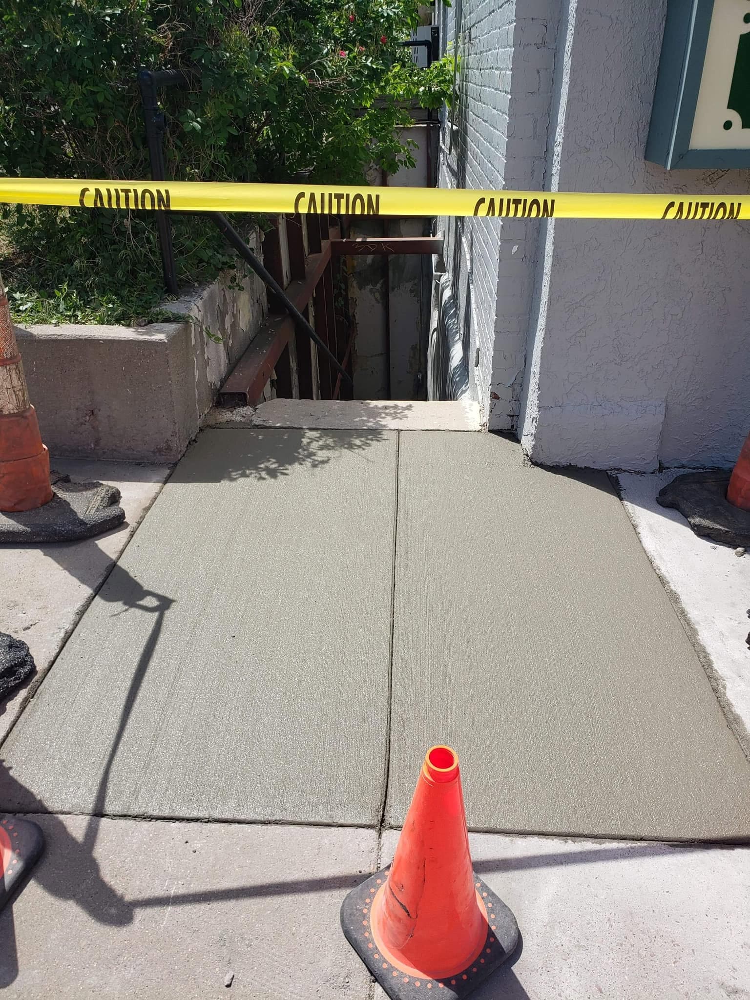 A fresh broom-finished concrete pad poured between a white brick storefront and a low concrete wall, taped off with caution tape and cones
