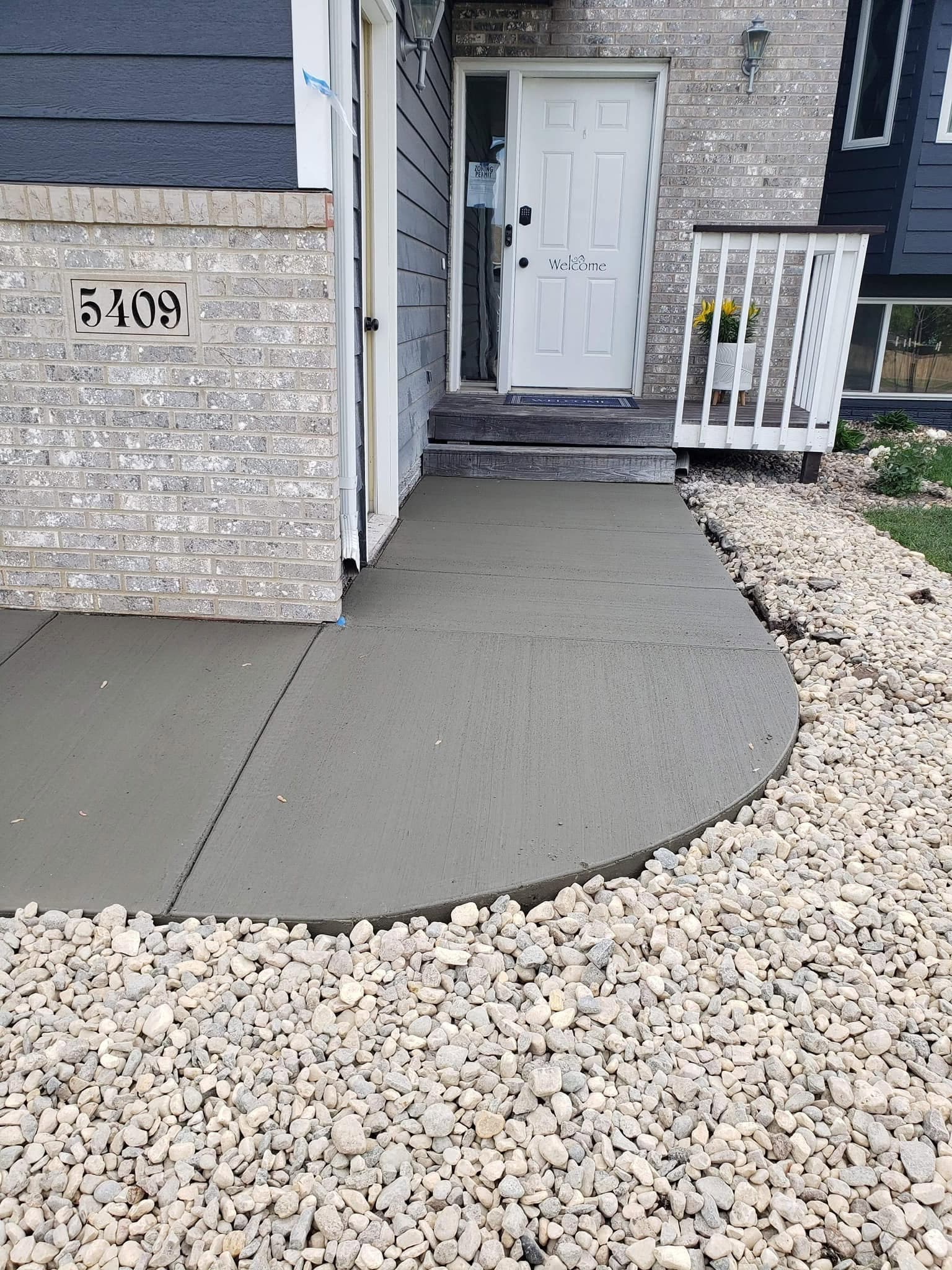 A fresh broom-finished concrete front walkway curving past a brick entry column to the front door, bordered with river rock
