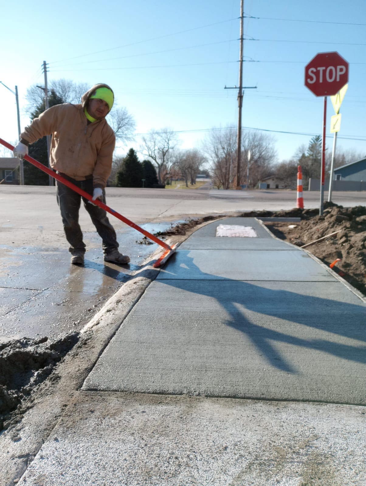 A worker dragging a broom finish across a fresh concrete sidewalk with a long-handled broom