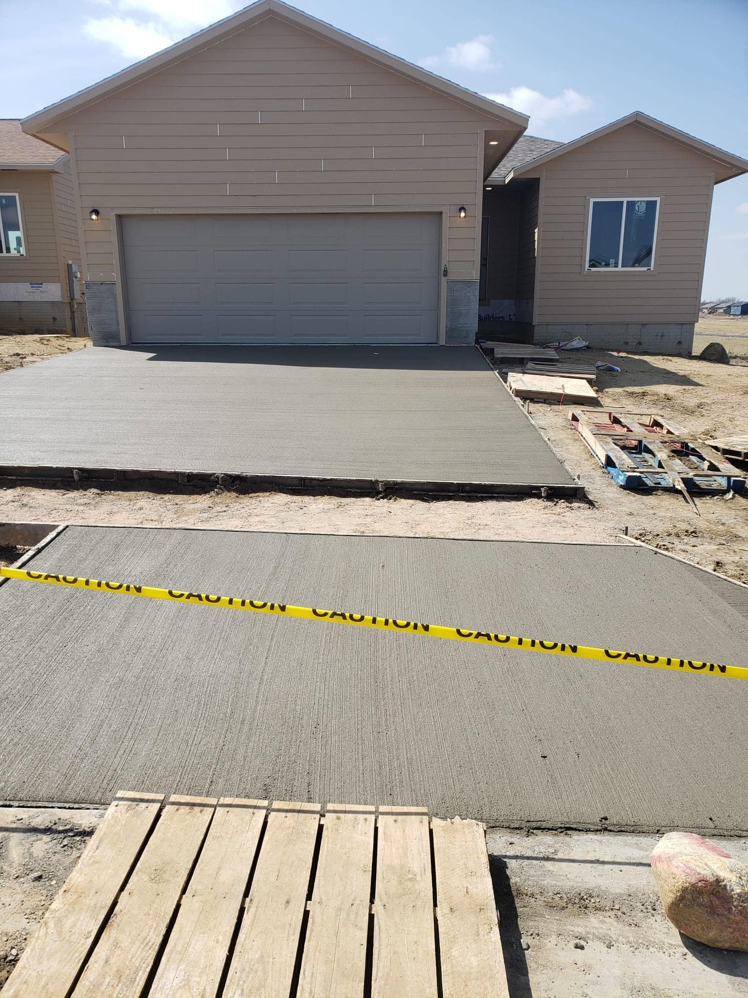 A fresh broom-finished concrete driveway curing behind caution tape in front of a new two-car garage, pallets and forms at its edges